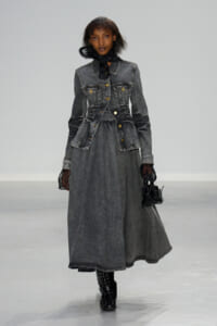 Model walking runway in a long gray denim dress with a matching fitted denim jacket, black sheer gloves, and a black scarf tied at the neck, carrying a small black handbag and wearing black textured boots.