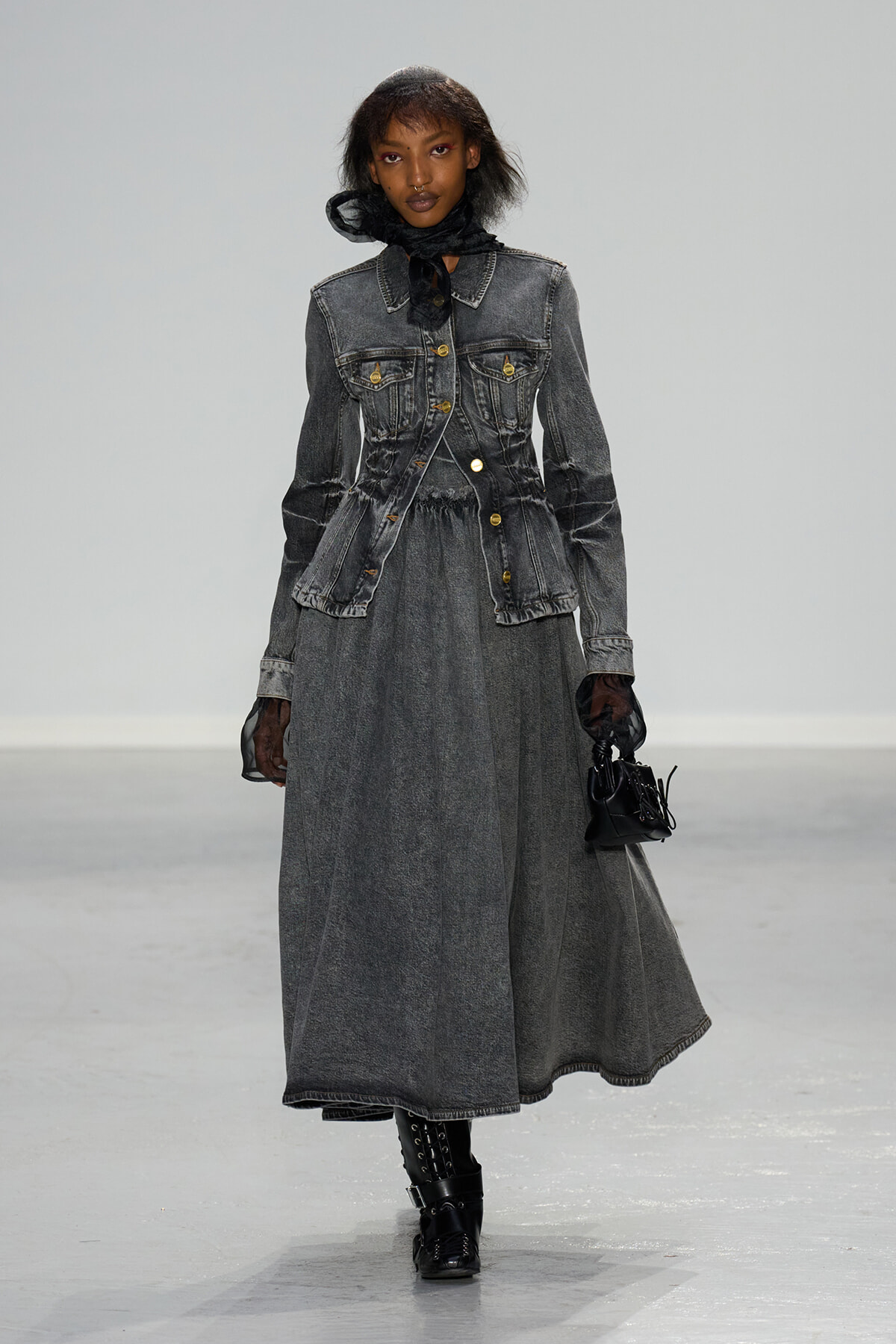 Model walking runway in a long gray denim dress with a matching fitted denim jacket, black sheer gloves, and a black scarf tied at the neck, carrying a small black handbag and wearing black textured boots.