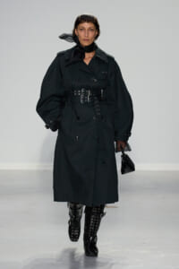 Model walking the runway in a black belted trench coat with oversized sleeves and knee-high studded boots.