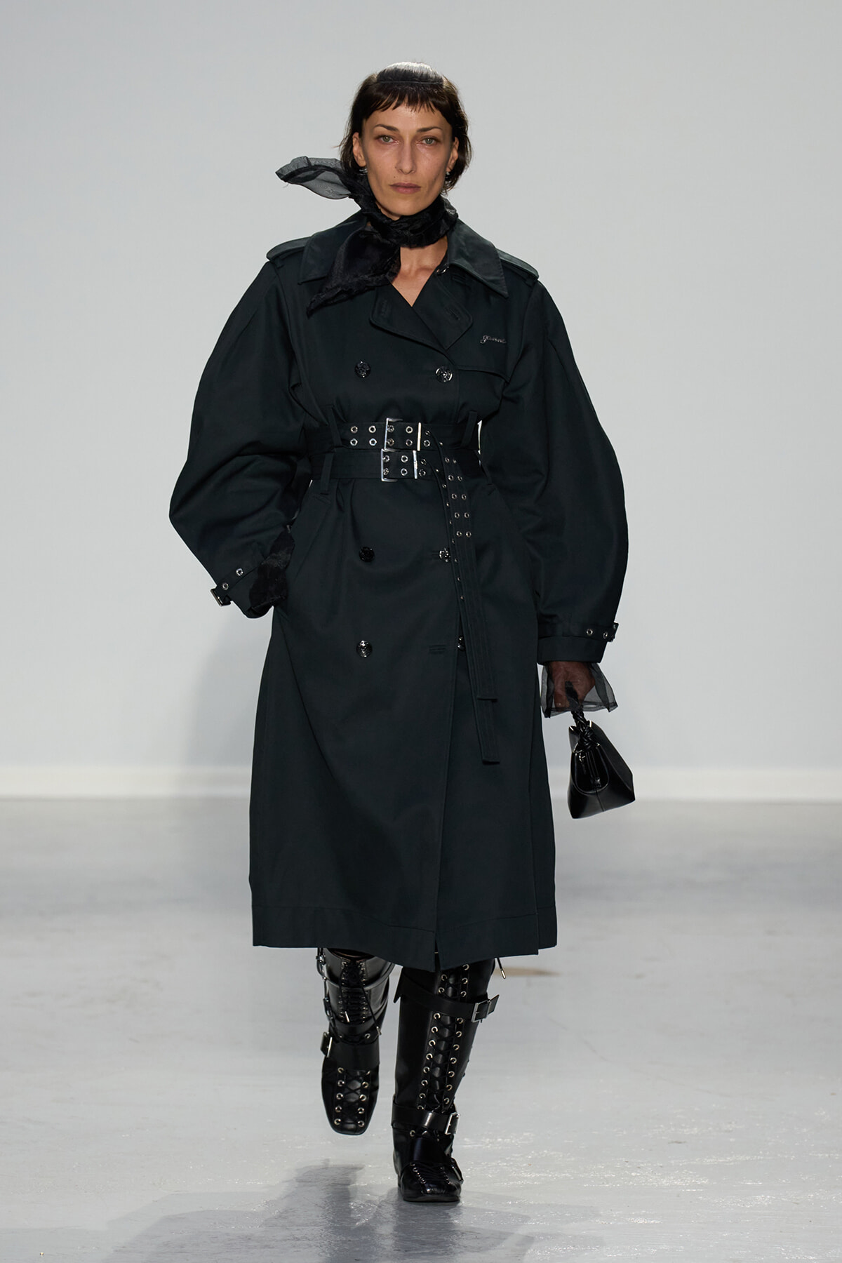 Model walking the runway in a black belted trench coat with oversized sleeves and knee-high studded boots.