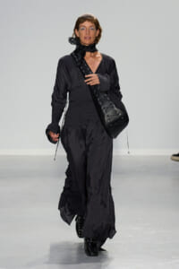 Female model walks a runway in an all-black, loose-fitting jumpsuit with a smocked waist and a large black lace-up crossbody bag worn over the shoulder.