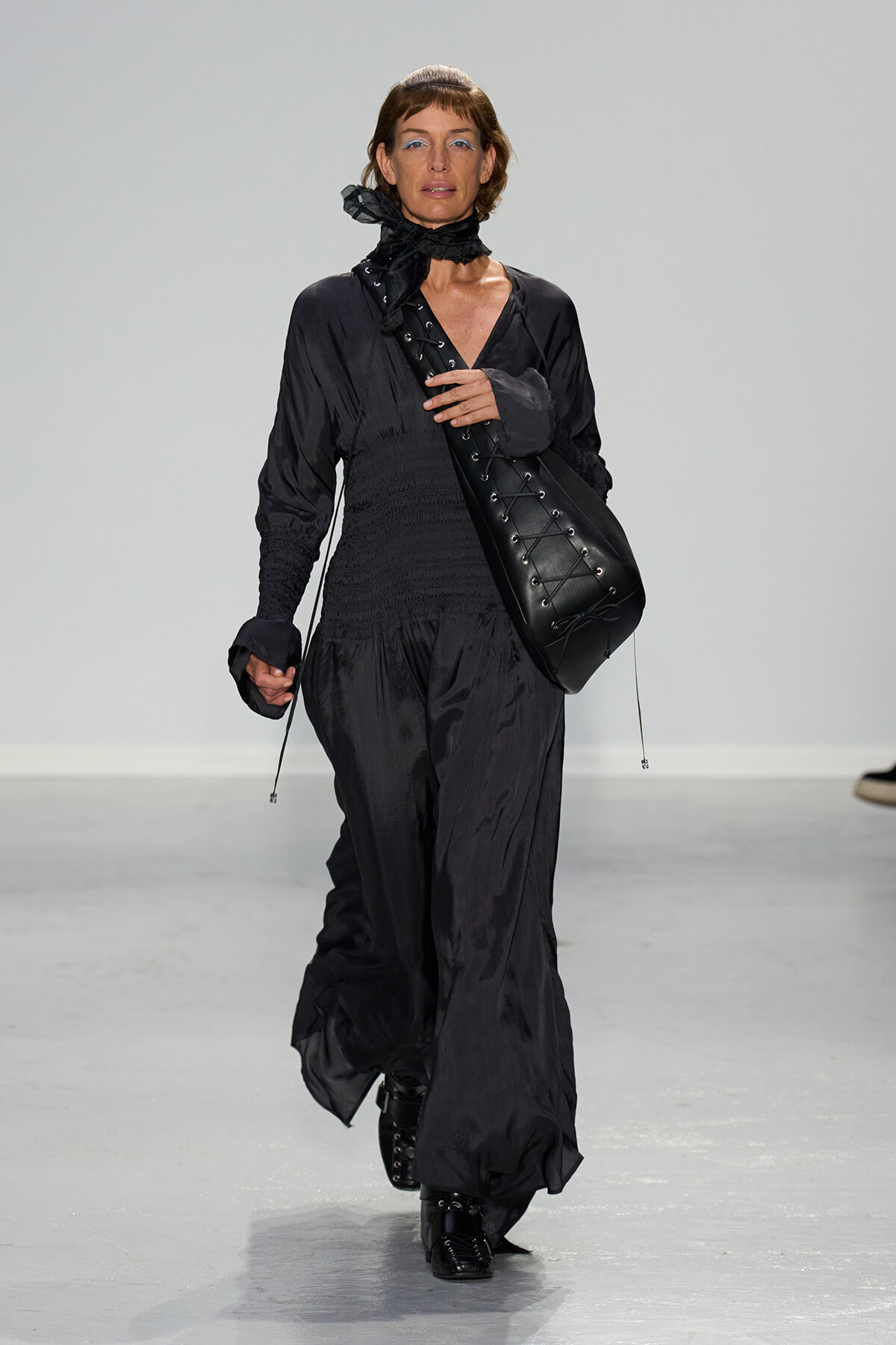 Female model walks a runway in an all-black, loose-fitting jumpsuit with a smocked waist and a large black lace-up crossbody bag worn over the shoulder.