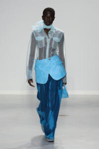 Runway model wearing a sheer light-blue button-down shirt with a ruffled scarf and a bright blue waist-wrap over wide-leg blue jeans, carrying a small blue handbag.