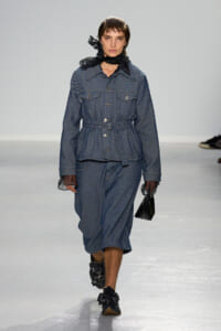 Model walks a runway in a blue pinstriped belted jacket and matching midi skirt, wearing a black scarf and sheer cuffs with floral-embellished shoes and a small black handbag.