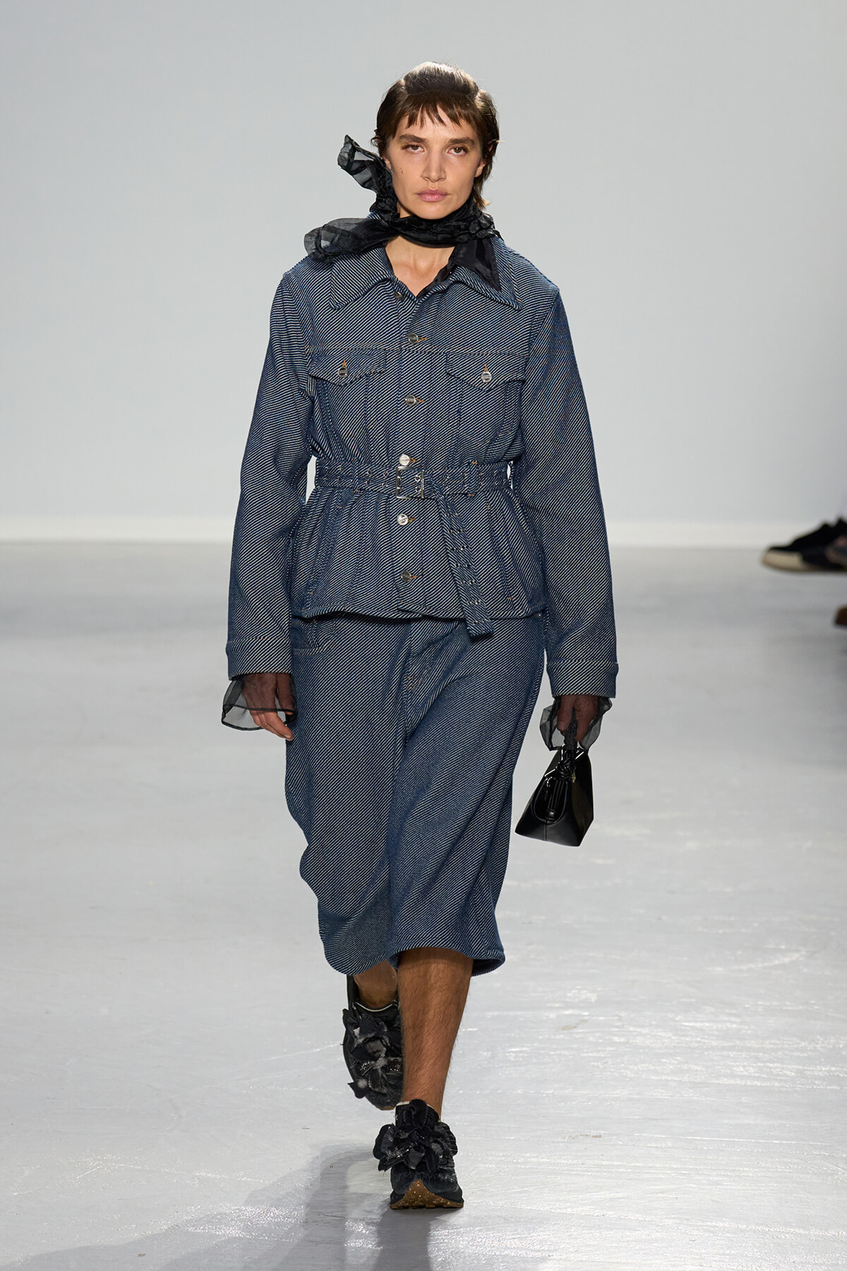 Model walks a runway in a blue pinstriped belted jacket and matching midi skirt, wearing a black scarf and sheer cuffs with floral-embellished shoes and a small black handbag.
