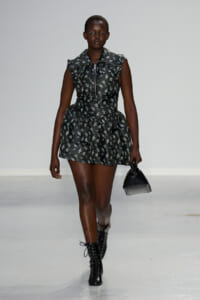 Model walking a runway in a black floral mini dress with a front zipper and ruffled sleeves, paired with black lace-up boots and a small black handbag.