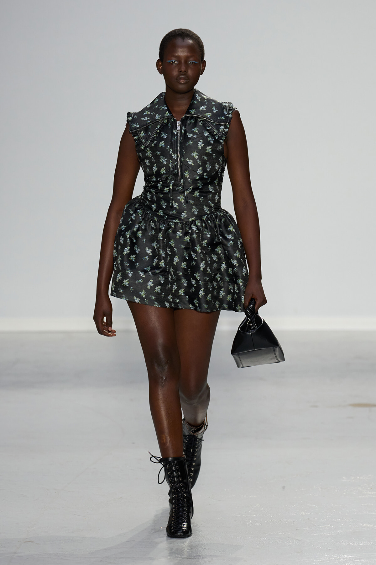 Model walking a runway in a black floral mini dress with a front zipper and ruffled sleeves, paired with black lace-up boots and a small black handbag.
