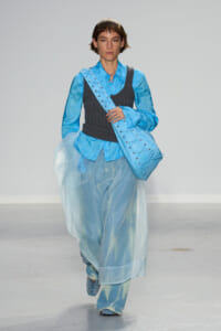 Model on a runway wearing layered blue garments: sheer skirt over pants, bright blue shirt, dark gray vest, and a large blue lace-up bag over the shoulder.