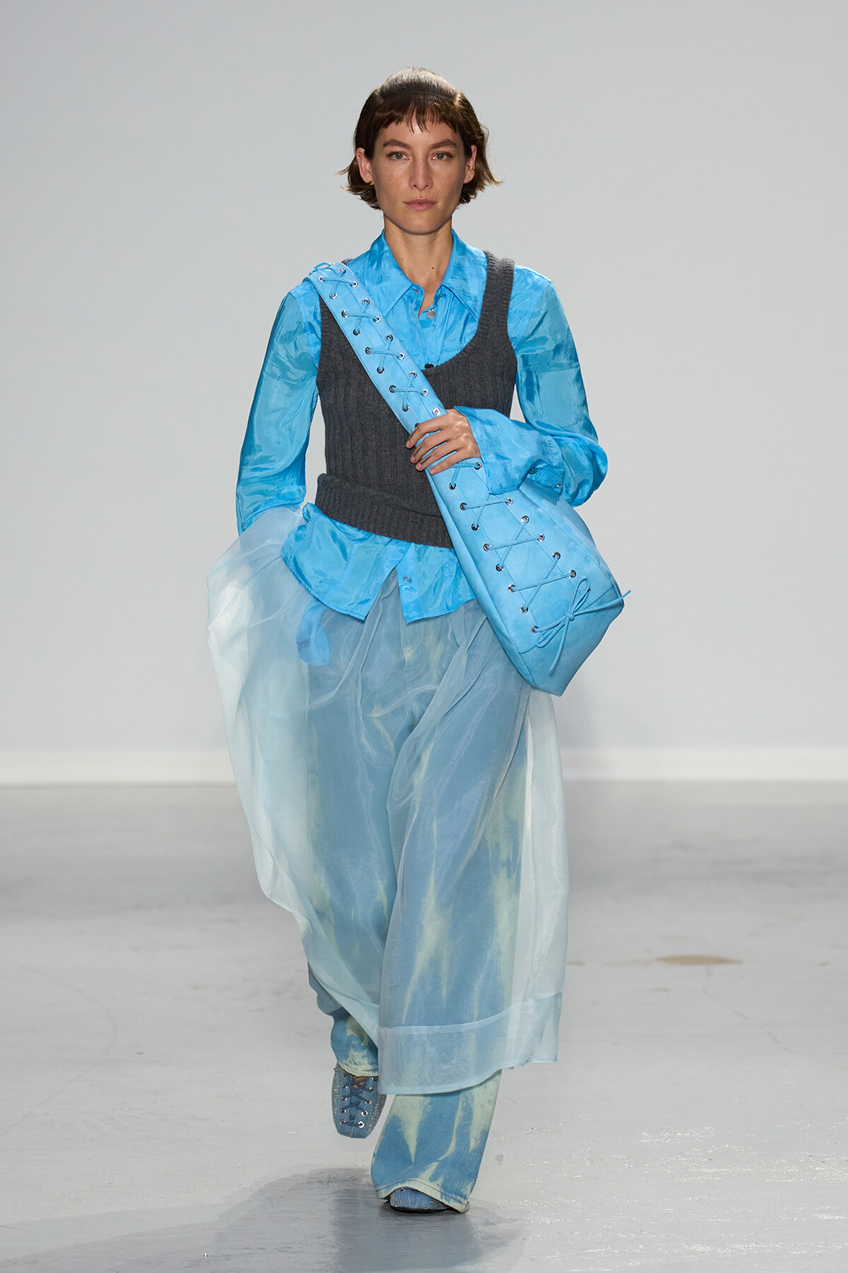 Model on a runway wearing layered blue garments: sheer skirt over pants, bright blue shirt, dark gray vest, and a large blue lace-up bag over the shoulder.