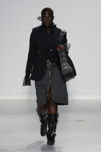Model walks a runway in a black oversized blazer and layered dark outfit, carrying a large black lace-up leather tote and wearing tall lace-up boots.