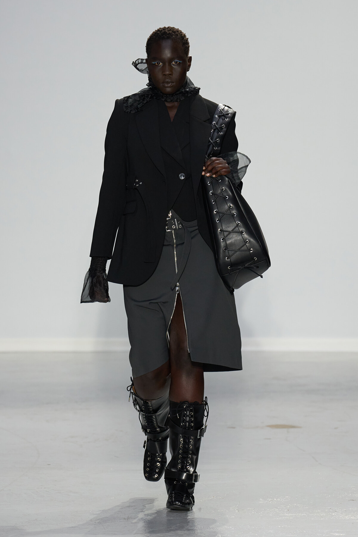 Model walks a runway in a black oversized blazer and layered dark outfit, carrying a large black lace-up leather tote and wearing tall lace-up boots.
