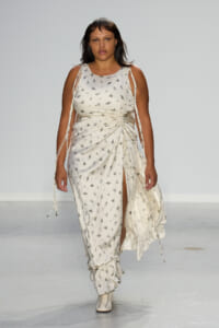Model walking the runway in a white floral print gown with ruching at the waist and a high slit.