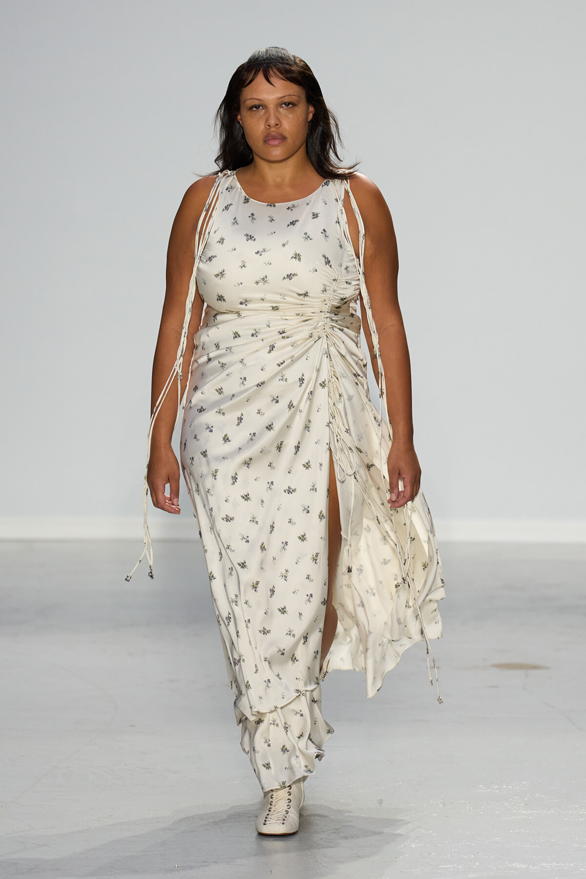 Model walking the runway in a white floral print gown with ruching at the waist and a high slit.