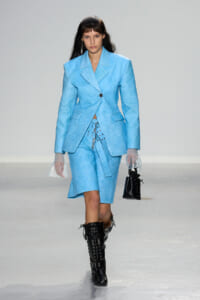 Model walking the runway in a light blue blazer and matching knee-length skirt, paired with black lace-up boots and sheer gloves.