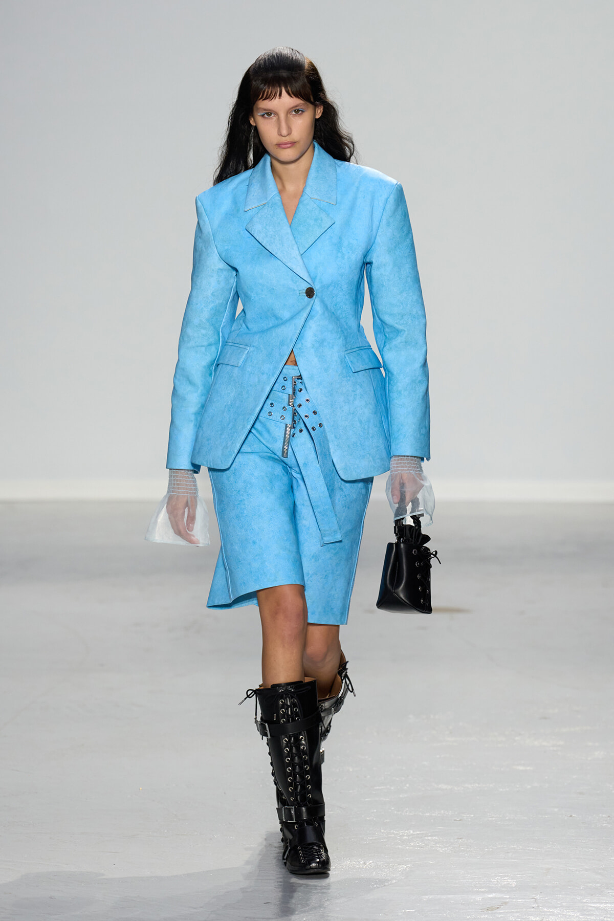Model walking the runway in a light blue blazer and matching knee-length skirt, paired with black lace-up boots and sheer gloves.