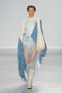 Model walks the runway in a pale blue sheer dress with a corseted bodice, paired with a chunky blue knit cardigan and thigh-high white lace-up boots, carrying a large white bag.