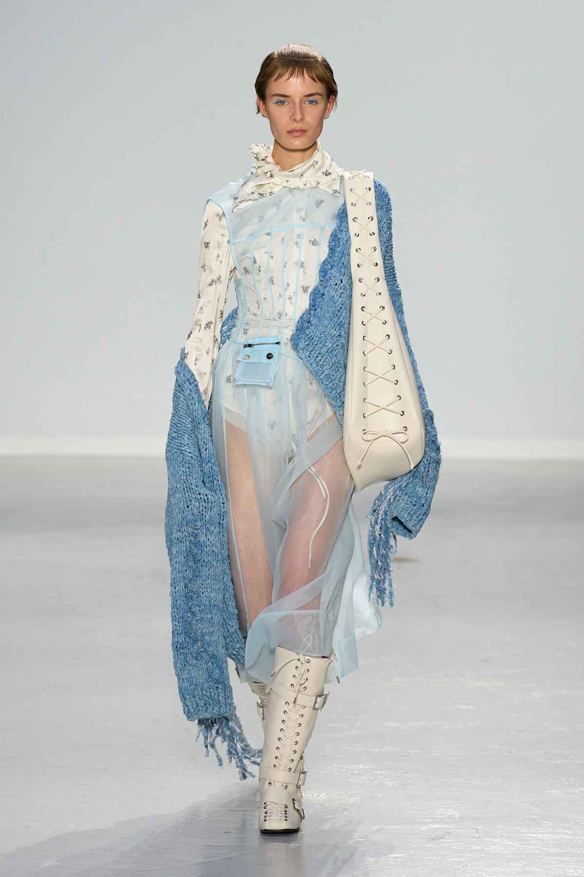 Model walks the runway in a pale blue sheer dress with a corseted bodice, paired with a chunky blue knit cardigan and thigh-high white lace-up boots, carrying a large white bag.