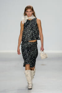 Model walking a runway in a black floral sleeveless top with matching floral shorts, a sheer white scarf, and cream lace-up knee-high boots, carrying a small white bag.