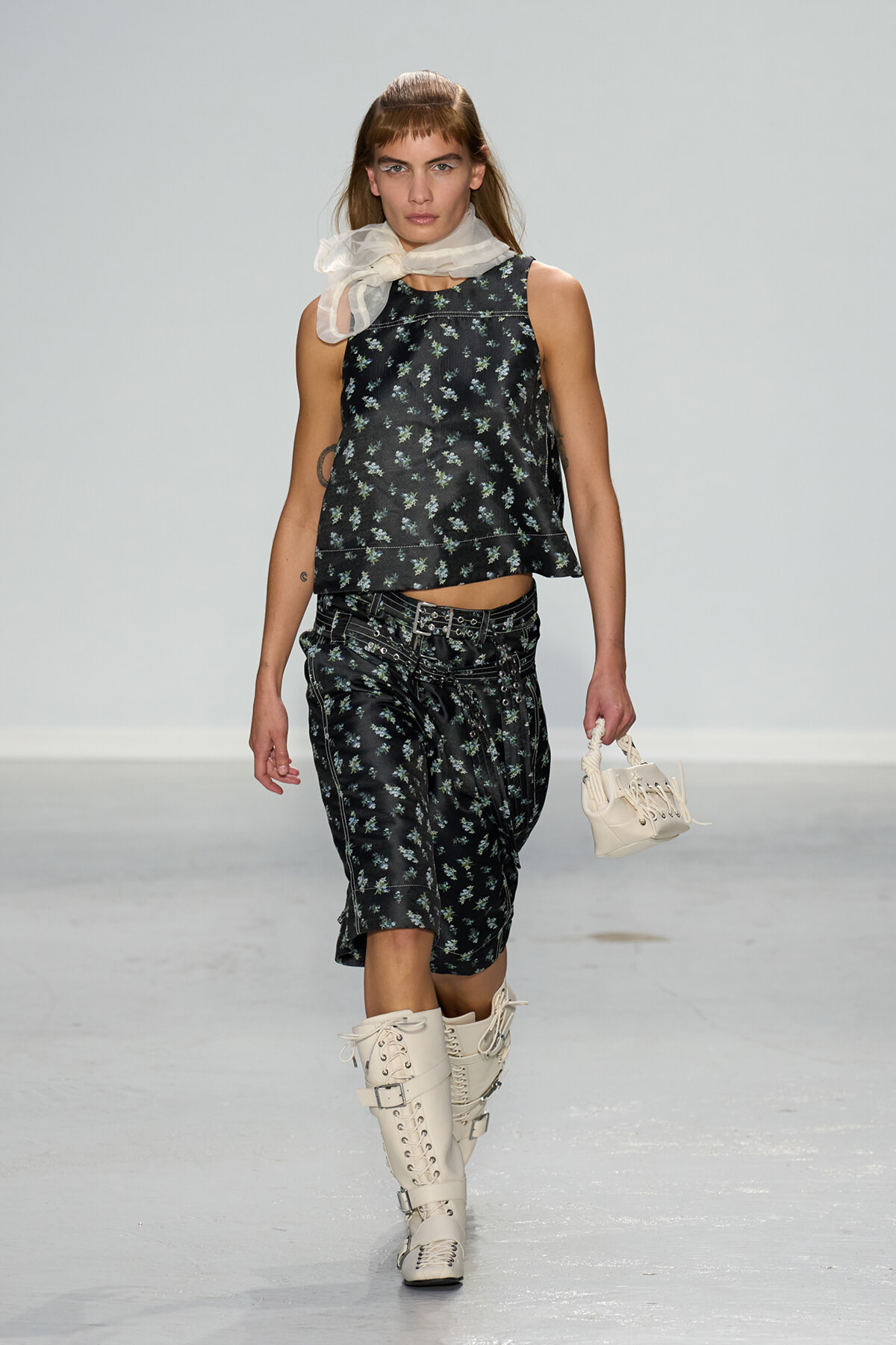 Model walking a runway in a black floral sleeveless top with matching floral shorts, a sheer white scarf, and cream lace-up knee-high boots, carrying a small white bag.