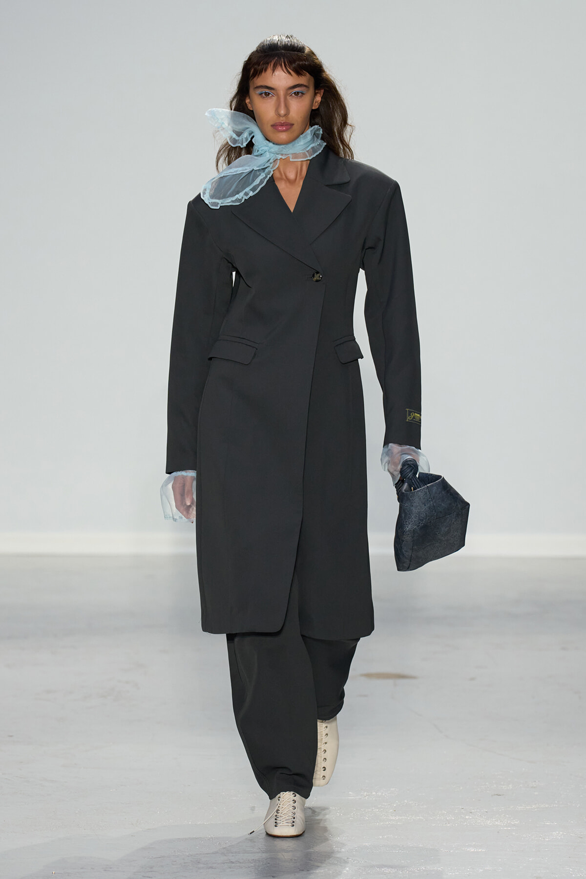 Model walks a fashion runway wearing a long dark blazer-style coat, light blue sheer scarf around the neck, cream lace-up boots, and a dark handbag.