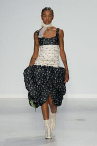 Model walking a runway in a black floral dress with a cream ruched waist, large gathered skirt, and white lace-up boots. Neck scarf adds to the look.