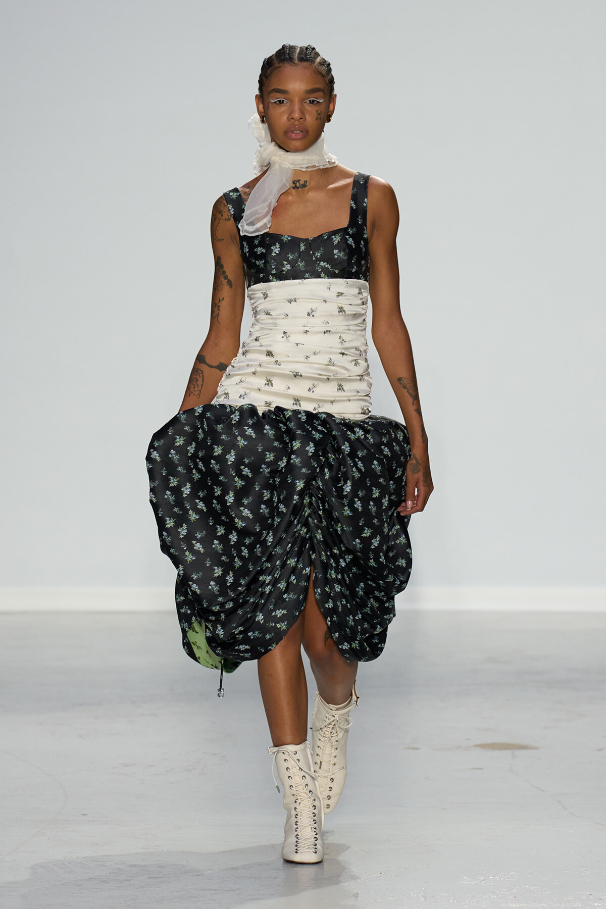 Model walking a runway in a black floral dress with a cream ruched waist, large gathered skirt, and white lace-up boots. Neck scarf adds to the look.