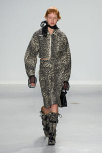 Model walks the runway in a leopard-print matching jacket and skirt set with a belt, black sheer gloves, and lace-up boots on a minimalist catwalk.