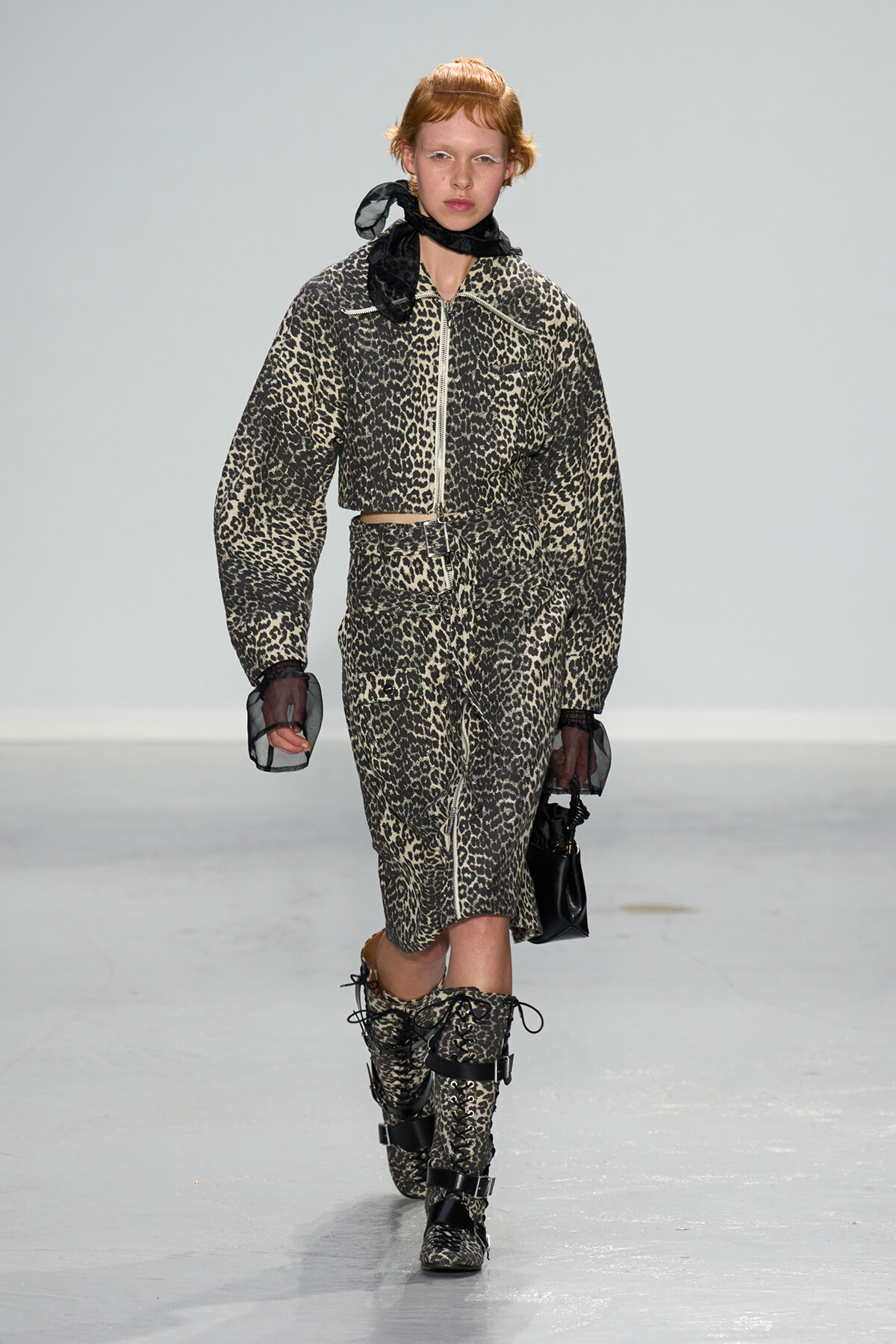 Model walks the runway in a leopard-print matching jacket and skirt set with a belt, black sheer gloves, and lace-up boots on a minimalist catwalk.