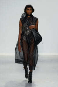 Model walks the runway in a sheer black ensemble with a sequined top and a large black lace-up shoulder bag over the shoulder.