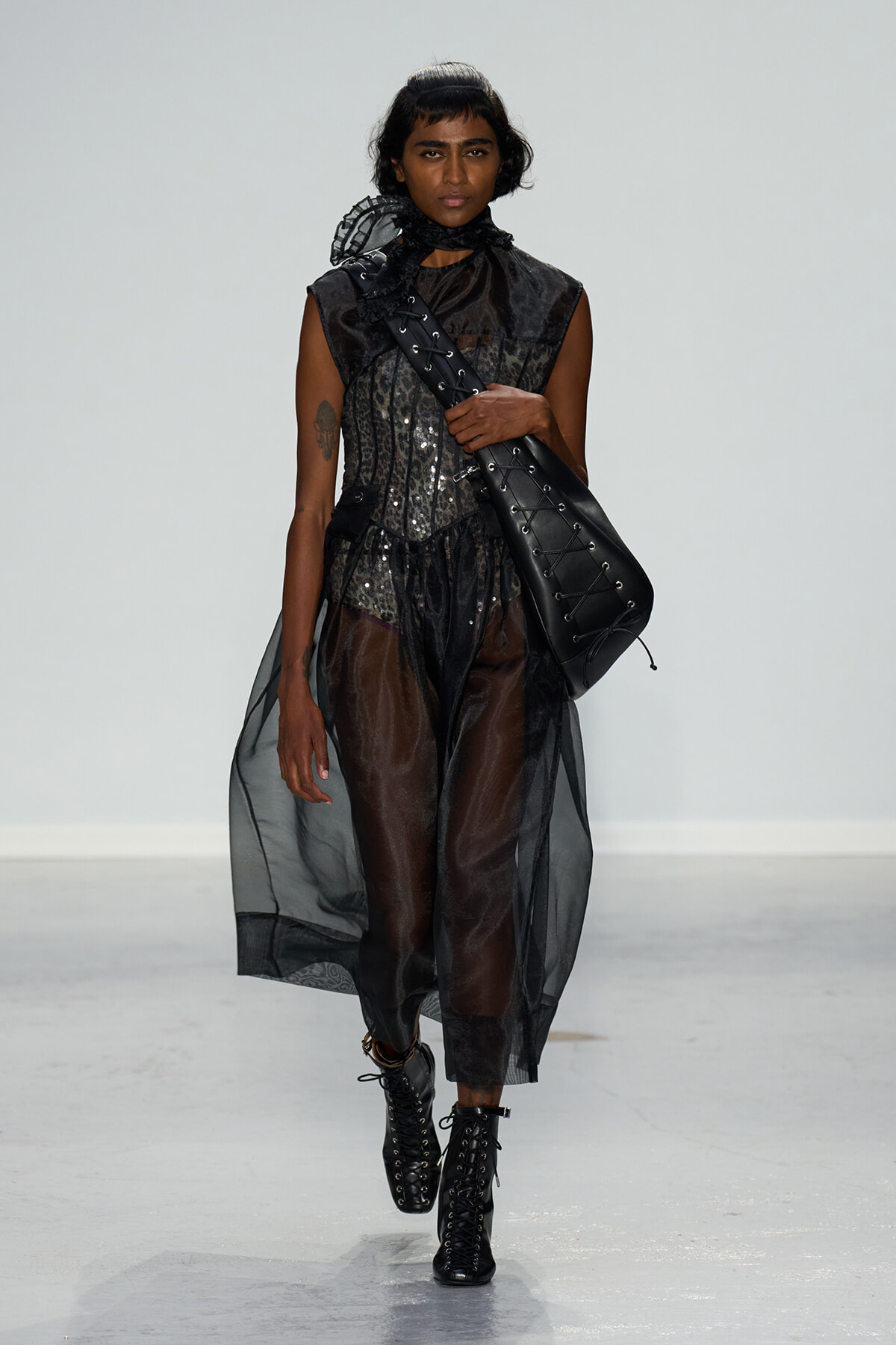 Model walks the runway in a sheer black ensemble with a sequined top and a large black lace-up shoulder bag over the shoulder.
