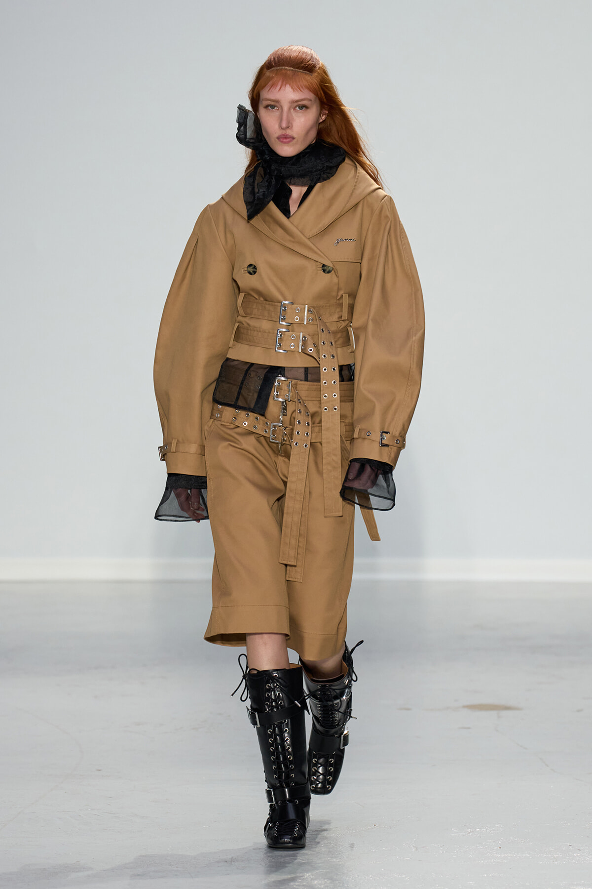 Model walking runway in tan belted trench ensemble with wide belts, black scarf, and chunky black lace-up boots.