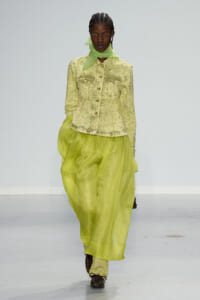 Model on a runway wearing lime-green sheer wide-leg pants and a pale embroidered denim jacket with a matching lime scarf.