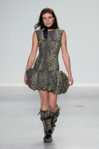 Model walking runway in a leopard-print sleeveless dress with a ruffled hem, black sheer neck scarf, and matching leopard boots with buckles.
