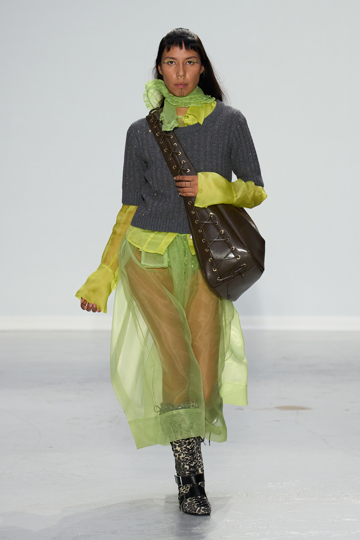 Runway model wearing a gray knit sweater with lime sheer layers, neon cuffs, a large brown lace-up shoulder bag, and leopard-print boots.