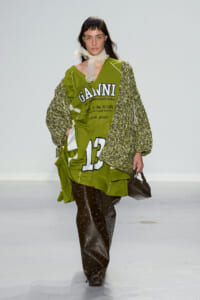 Model walks the runway in an oversized olive-green jersey dress with bold graphics and large white numbers, layered with a chunky knit cardigan and black studded pants, carrying a small black handbag.