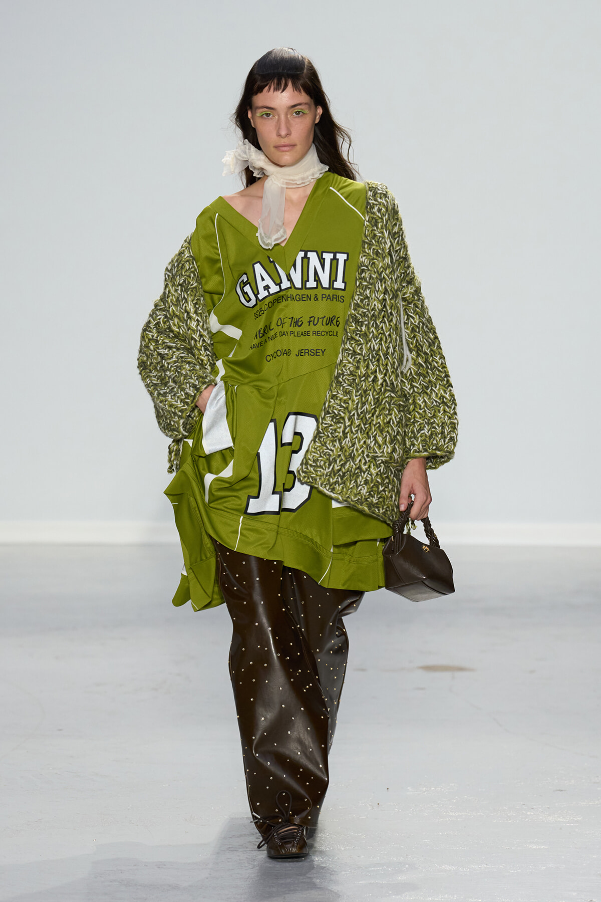 Model walks the runway in an oversized olive-green jersey dress with bold graphics and large white numbers, layered with a chunky knit cardigan and black studded pants, carrying a small black handbag.