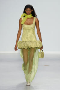 Model walking runway in a pale yellow-green dress with a voluminous bubble skirt and sheer matching pants, accessorized with a lime scarf.