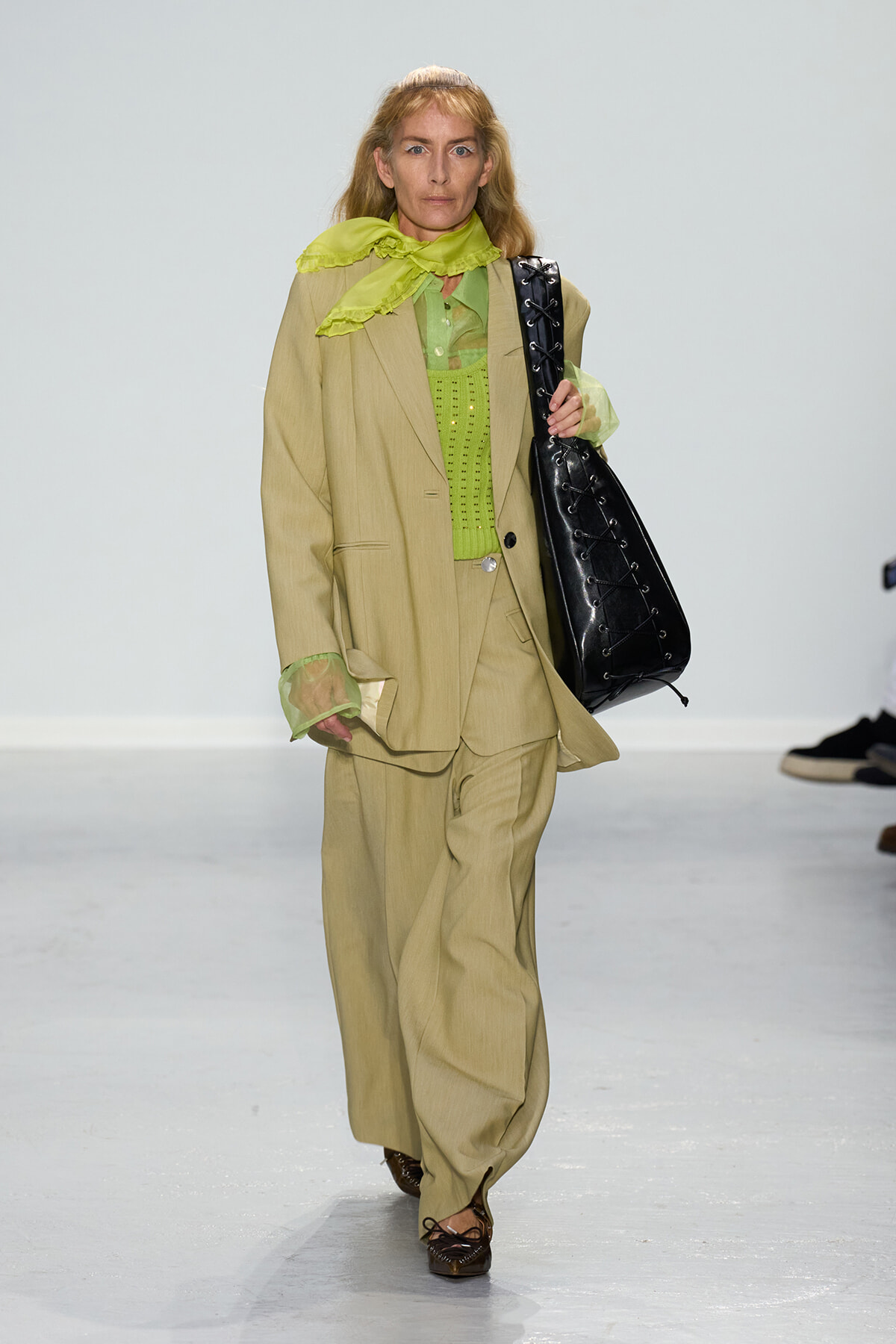 Model walking a fashion runway in a beige oversized suit, lime green sheer blouse and scarf, carrying a large black laced leather tote.