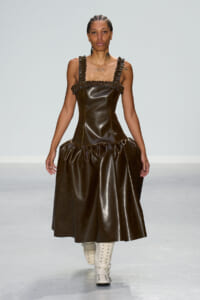 Runway model wearing a dark brown leather dress with ruffled straps and a voluminous skirt, paired with white lace-up boots.