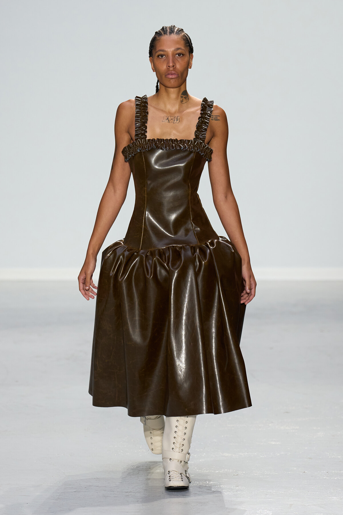 Runway model wearing a dark brown leather dress with ruffled straps and a voluminous skirt, paired with white lace-up boots.