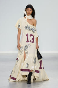 Model walking a runway in a cream, oversized one-shoulder dress with large purple '13' and scattered graphic text, paired with black lace-up boots and a small black handbag, scarf tied at the neck.