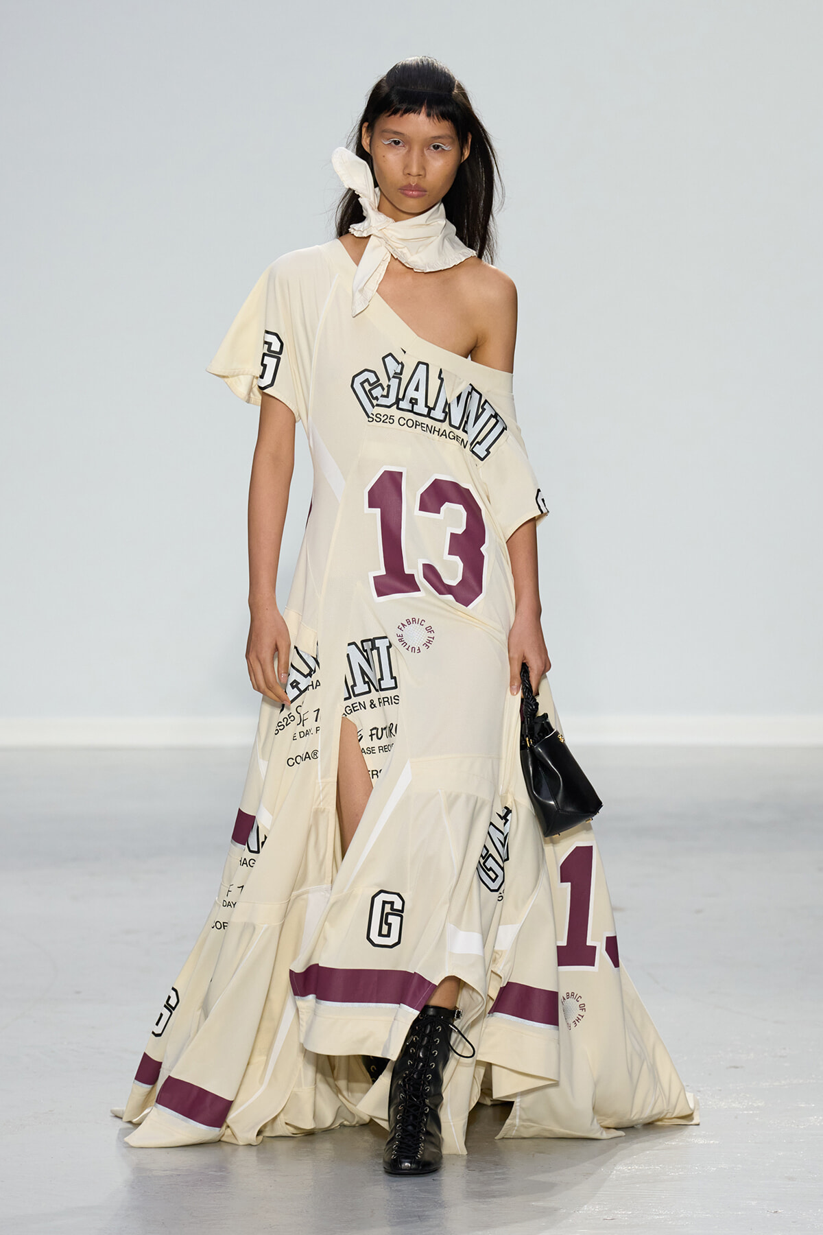 Model walking a runway in a cream, oversized one-shoulder dress with large purple '13' and scattered graphic text, paired with black lace-up boots and a small black handbag, scarf tied at the neck.