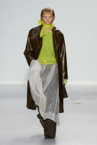 Model walking a runway in a bright lime knit top, sheer white skirt over dark pants, and a dark brown leather coat with a lime scarf.
