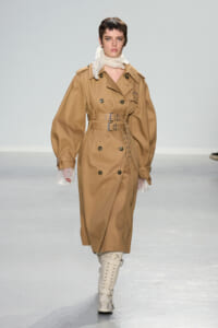 Model walking a runway in a tan trench coat with a wide belt, scarf at the neck, and cream knee-high lace-up boots.