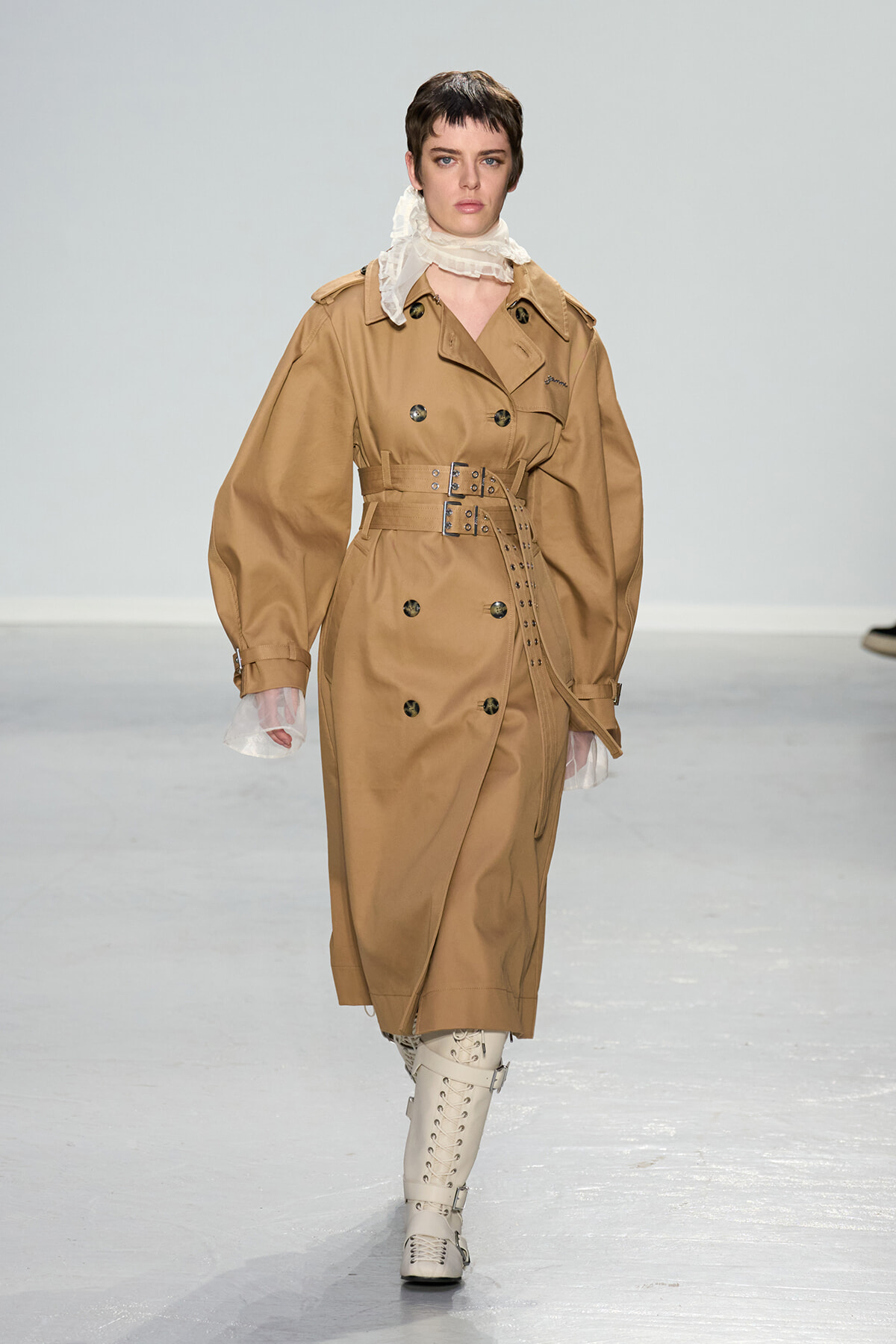Model walking a runway in a tan trench coat with a wide belt, scarf at the neck, and cream knee-high lace-up boots.
