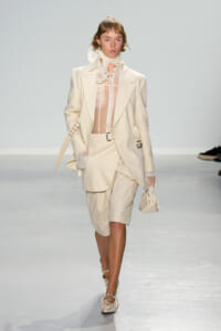 Model on a fashion runway wearing a cream suit with an oversized blazer, sheer blouse, and matching shorts, carrying a small cream handbag.