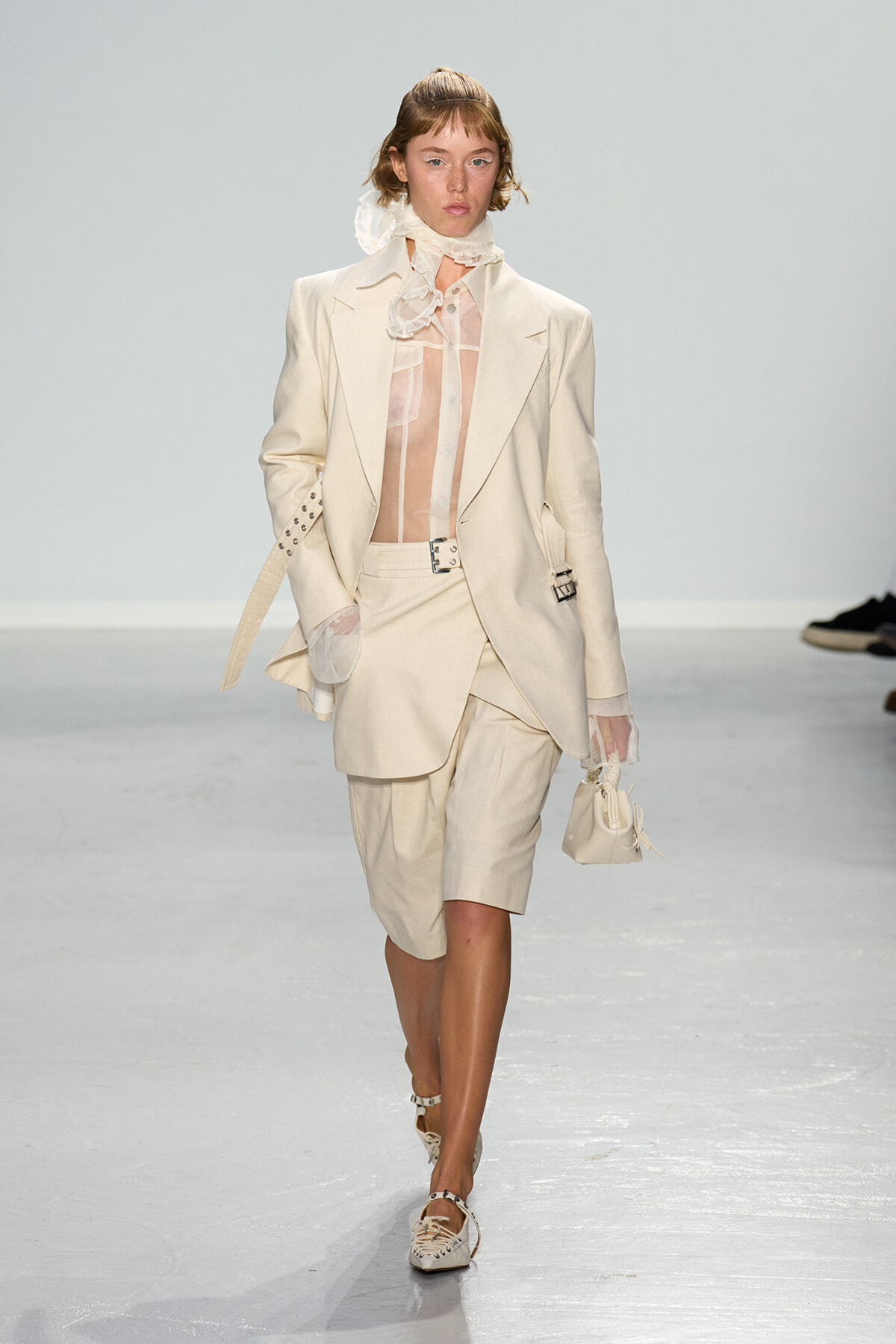 Model on a fashion runway wearing a cream suit with an oversized blazer, sheer blouse, and matching shorts, carrying a small cream handbag.