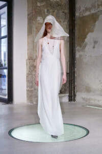 Model in a flowing white sleeveless dress with a large translucent white hat, standing on a circular glass platform in an industrial-styled studio.