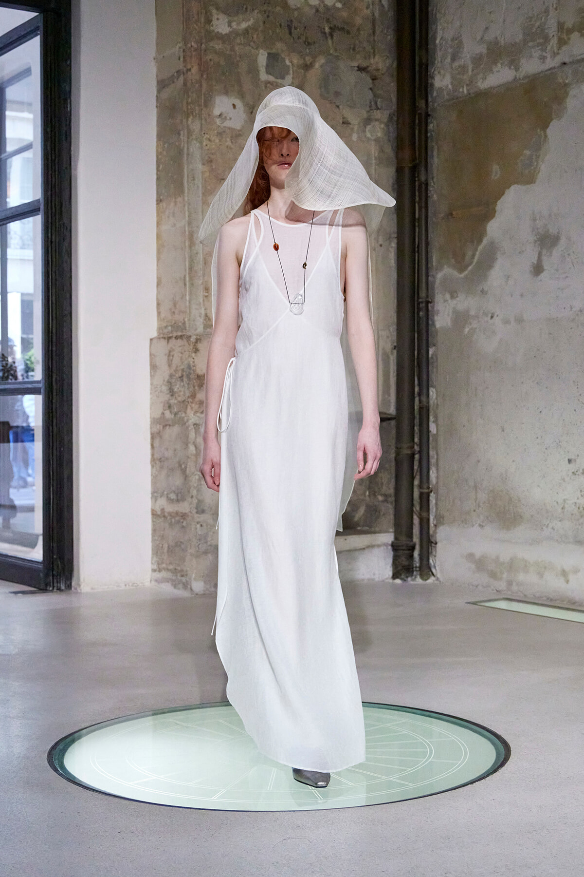 Model in a flowing white sleeveless dress with a large translucent white hat, standing on a circular glass platform in an industrial-styled studio.
