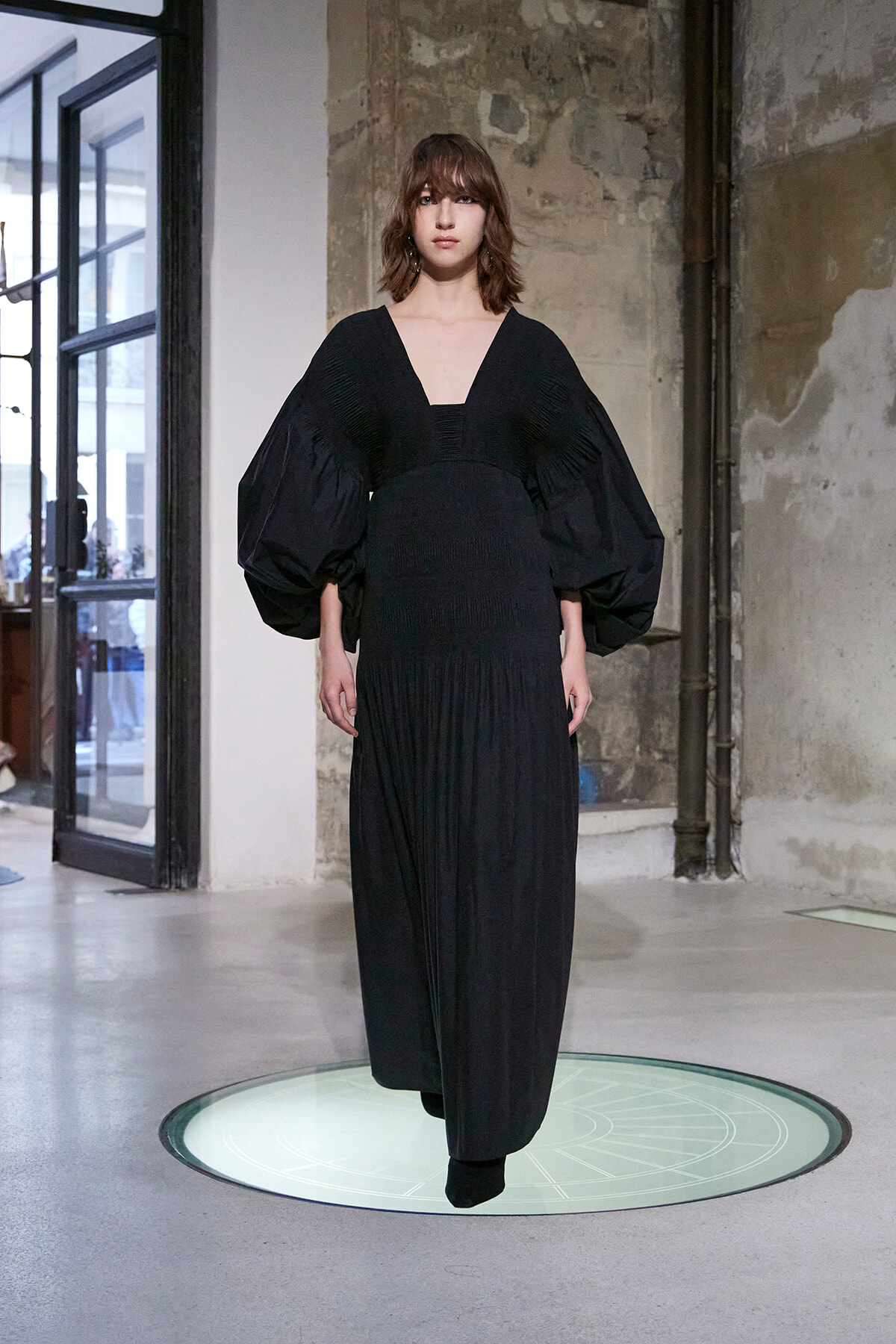 Model in a black, floor-length dress with oversized puff sleeves on a circular light platform.